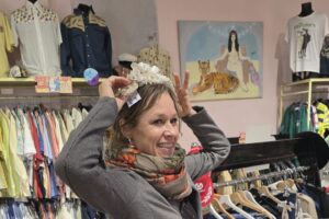 A smiling woman in a grey cardigan and patterned scarf tries on an ornate headpiece beside clothing racks in a vintage shop.